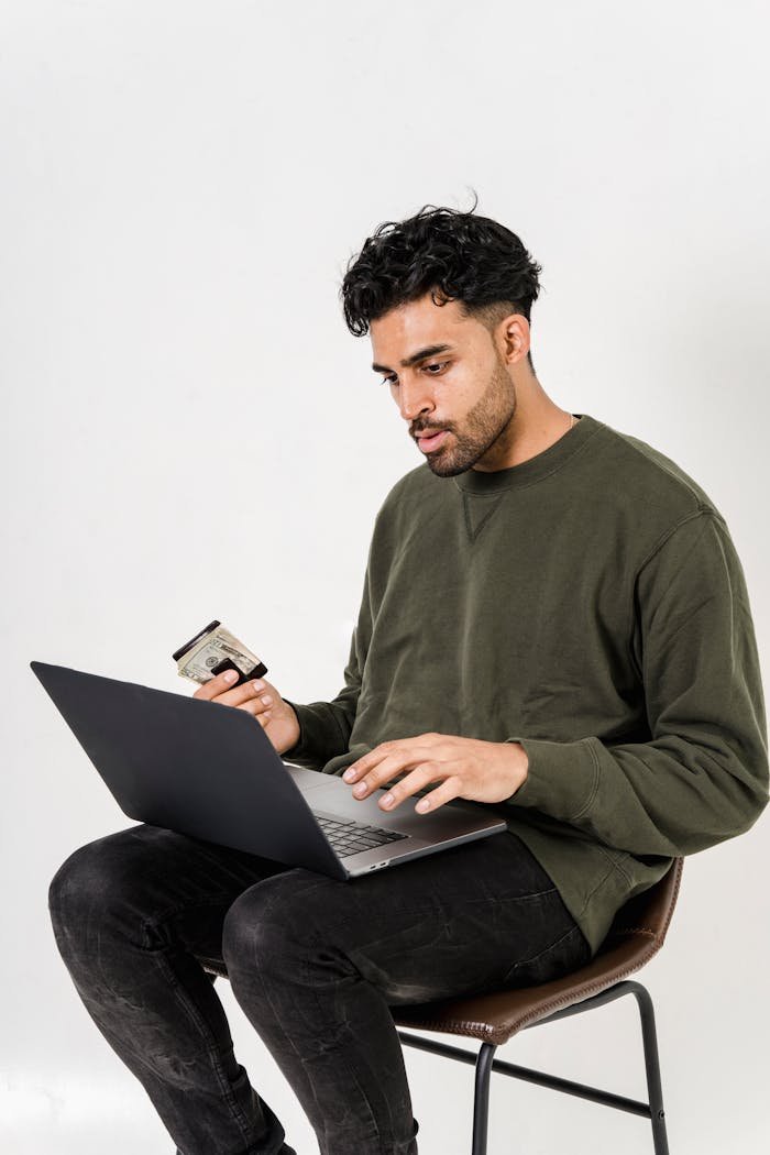 Young man sitting indoors shopping online using a laptop and credit card, showcasing modern technology and convenience.